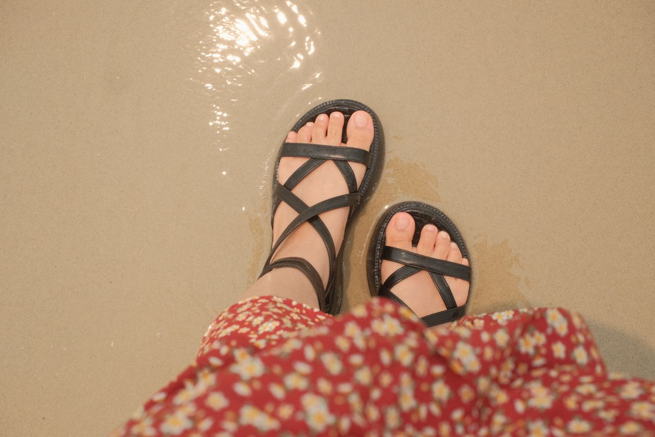 services-img Top view of woman's feet in black sandals on a sandy beach, wearing a floral dress.
