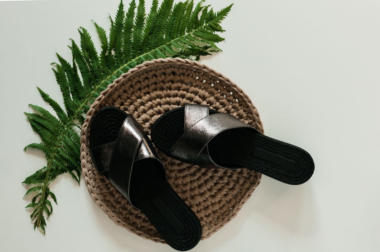 gallery-1 Pair of elegant black sandals placed on a woven mat with a decorative fern leaf.